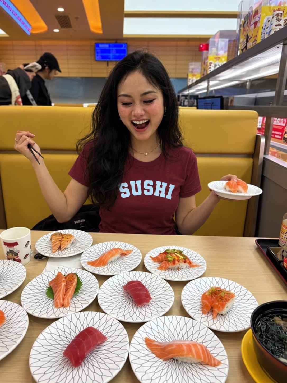 A woman sitting in a sushi restaurant wearing a maroon t-shirt with 'SUSHI' printed in white. She is holding chopsticks and a small plate of sushi, with multiple plates of sushi spread out. sushi varsity tshirt on the table in front of her.