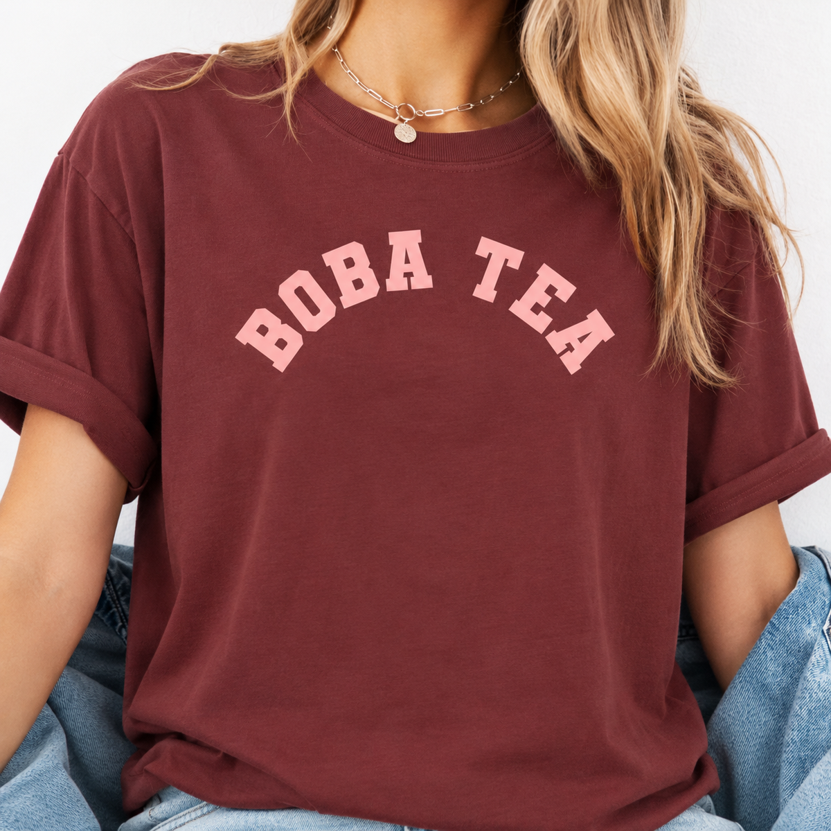A woman wearing a maroon short-sleeve t-shirt with 'BOBA TEA' printed in light pink arched letters across the chest, paired with light blue jeans. Boba tea slogan T-shirt for fans of playful foodie fashion