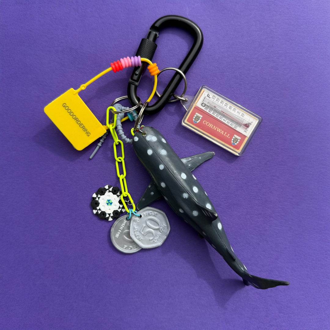 A shark-shaped keychain with a black carabiner, yellow and neon chain, coin charms, a small card, and a yellow tag, displayed on a purple background.
