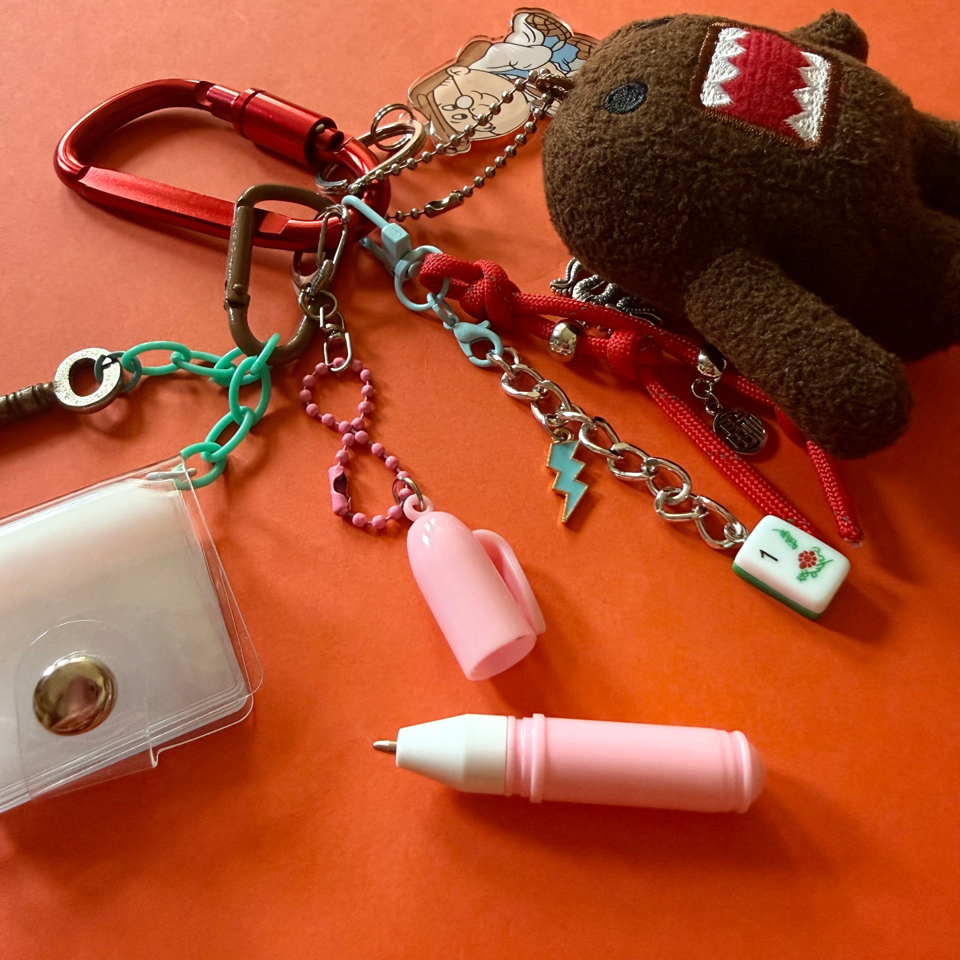 A keychain organizer set featuring a brown plush character, a red carabiner, a pink lip balm, a clear card holder, and several colorful charms on a bright orange background.