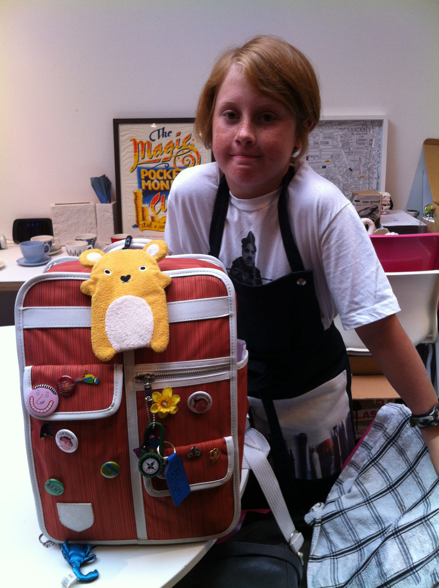 A child wearing a white t-shirt and black apron stands behind a red striped fabric suitcase decorated with buttons, patches, and a yellow plush toy, in an indoor room setting.
