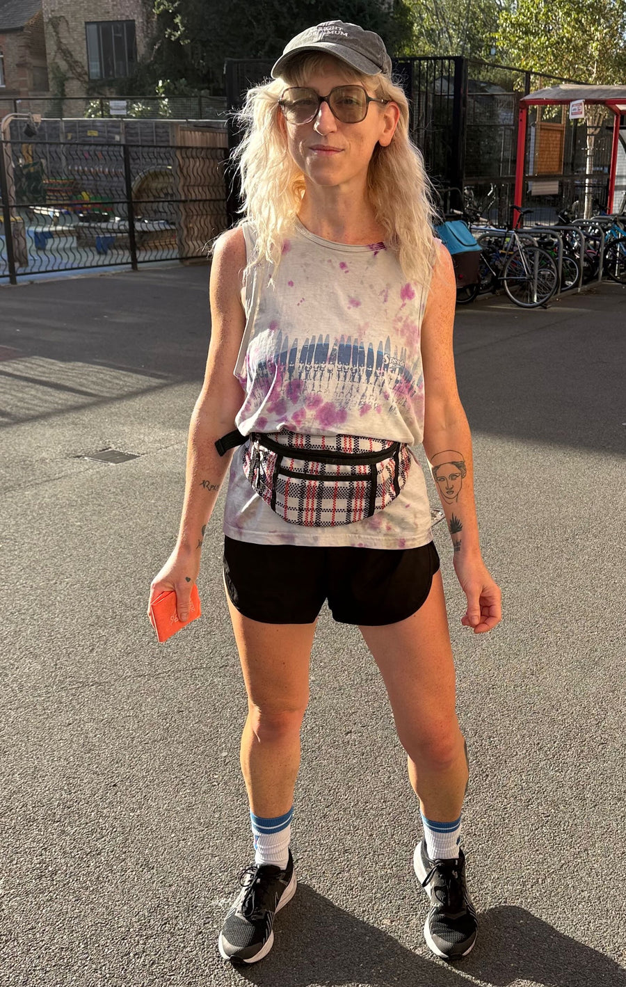 A woman standing outdoors on a paved area wearing a sleeveless tie-dye tank top, black shorts, a gray cap, sunglasses, white socks, and black sneakers. She has a plaid fanny pack around her waist and is holding an orange item in one hand. Goodordering bum bag