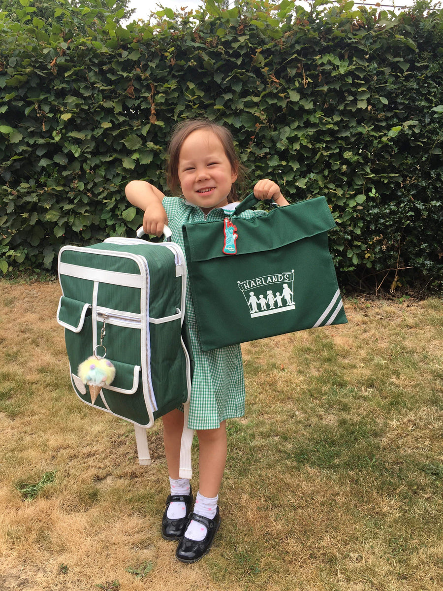 A young girl stands outdoors on grass holding two green fabric bags: a small backpack with white trim and a larger tote bag with a white printed logo. The background is a dense green hedge.