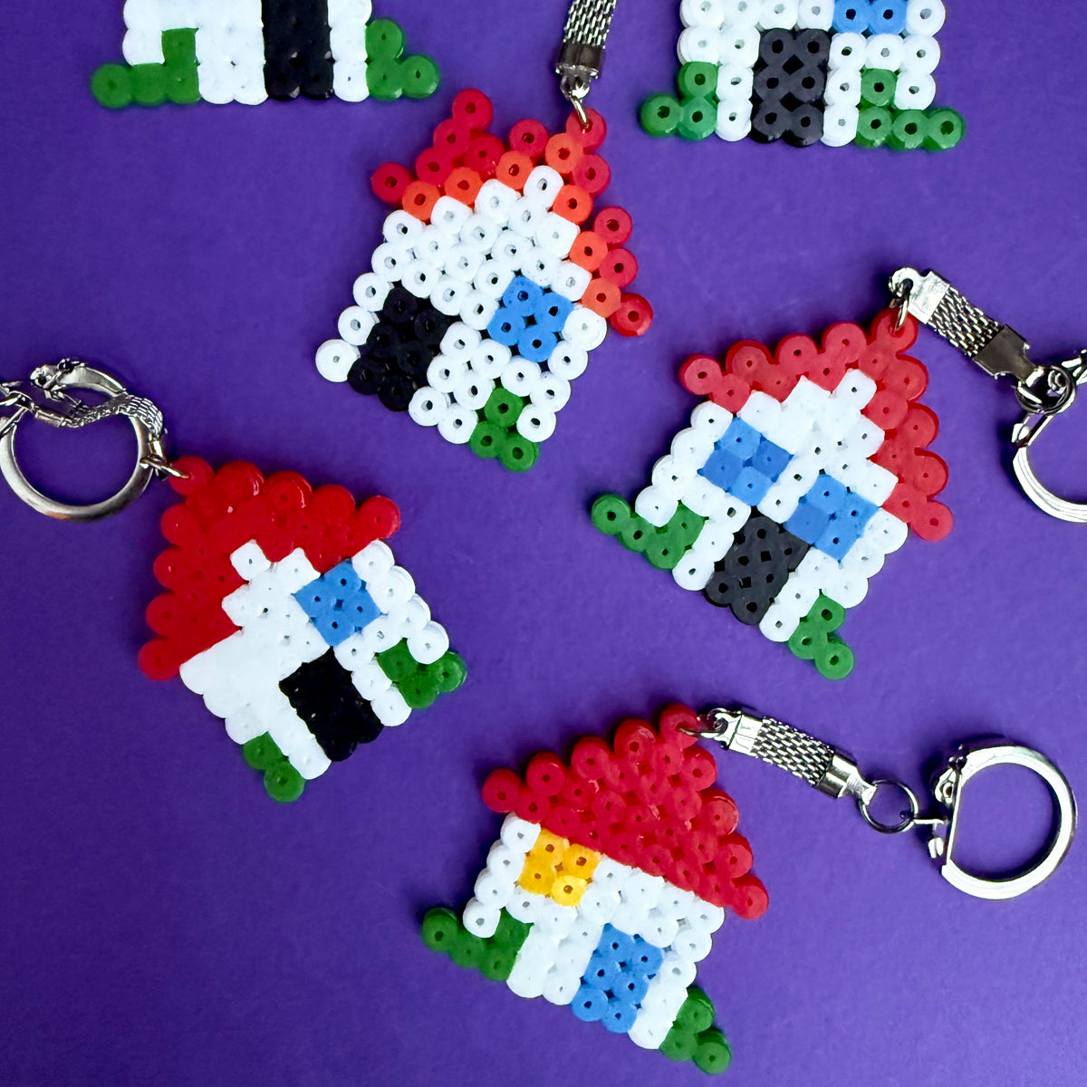 Several small house-shaped keychains made from colorful fused beads are arranged on a purple background. The bead houses use red, white, green, black, blue, and yellow colors, with silver metal key rings attached.
