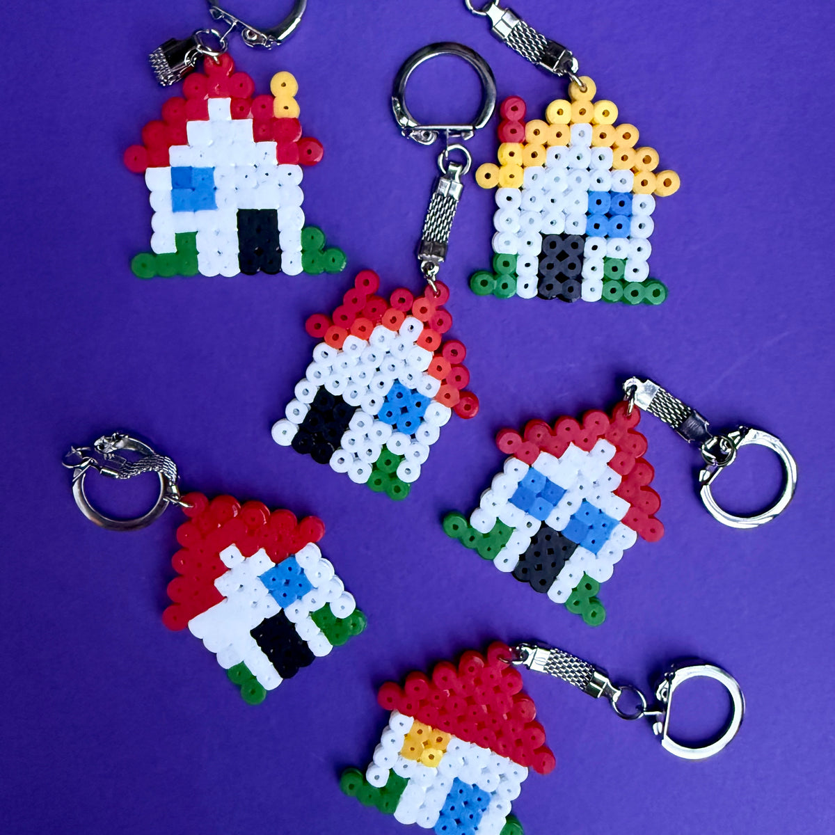 Six handmade bead keychains shaped like houses, arranged on a solid purple background. The keychains feature silver metal keyrings and are crafted from small colorful beads representing roofs, walls, windows, doors, and grass.
