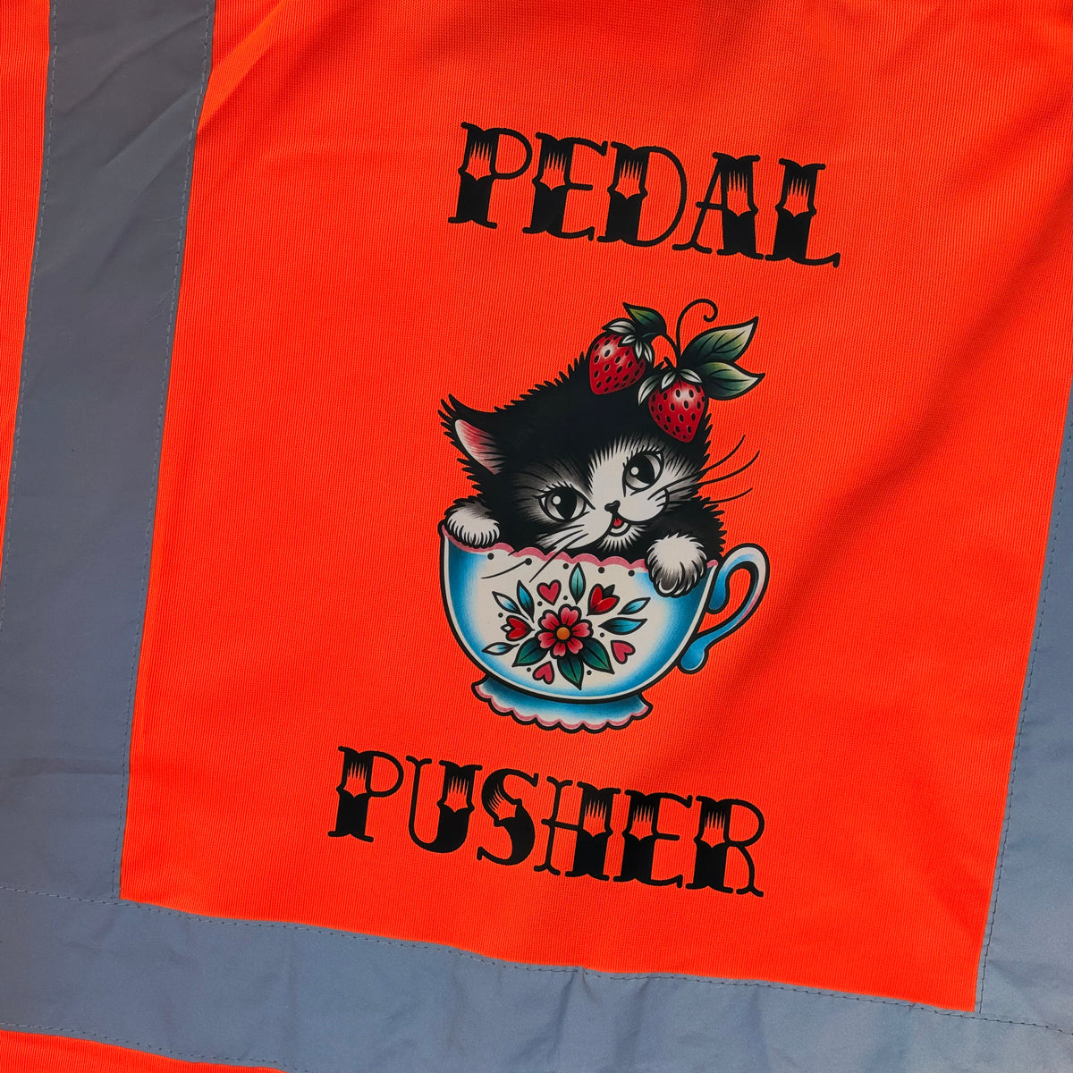 Close-up of a bright orange high vis cycling vest with  a central graphic showing a black-and-white kitten in a floral teacup with strawberries, surrounded by the words “PEDAL PUSHER” in black.