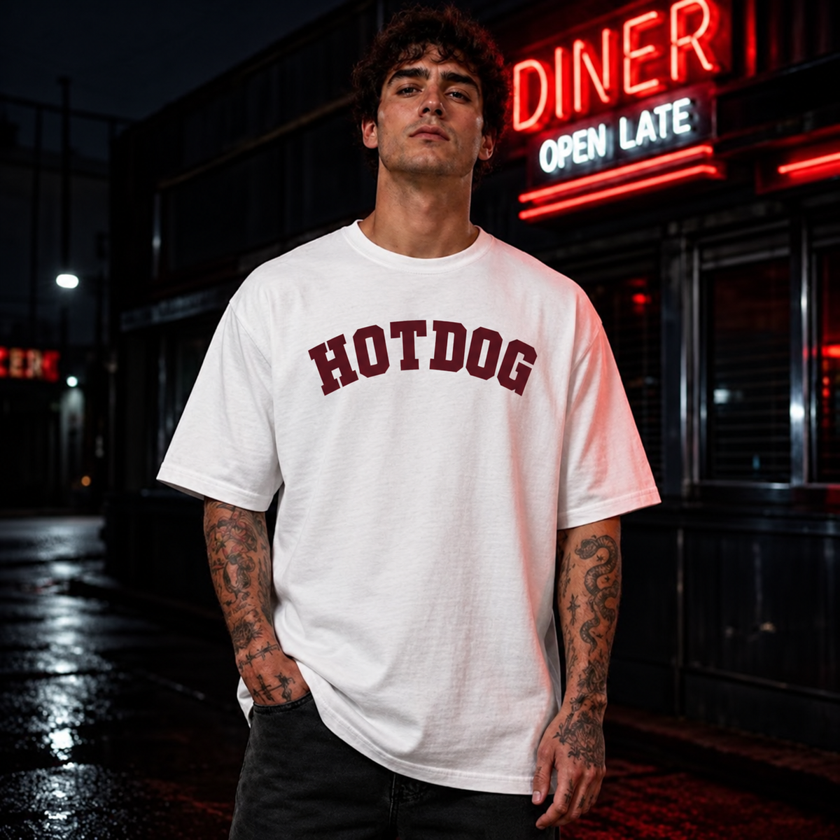 A man wearing a white t-shirt with 'HOTDOG' printed in maroon arched letters across the chest, standing in front of a dark night-time diner with a red neon sign. Hot dog graphic tee available in multiple colours