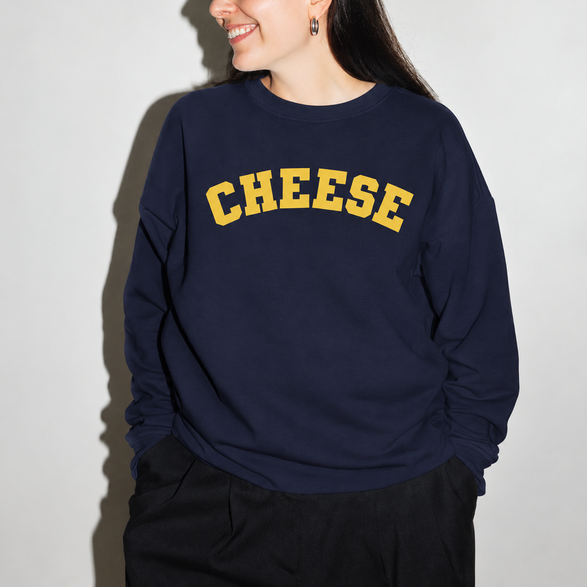 A woman wearing a navy blue crewneck sweatshirt with the word 'CHEESE' printed across the chest in yellow varsity-style lettering, paired with black pants. Food lover's gift
