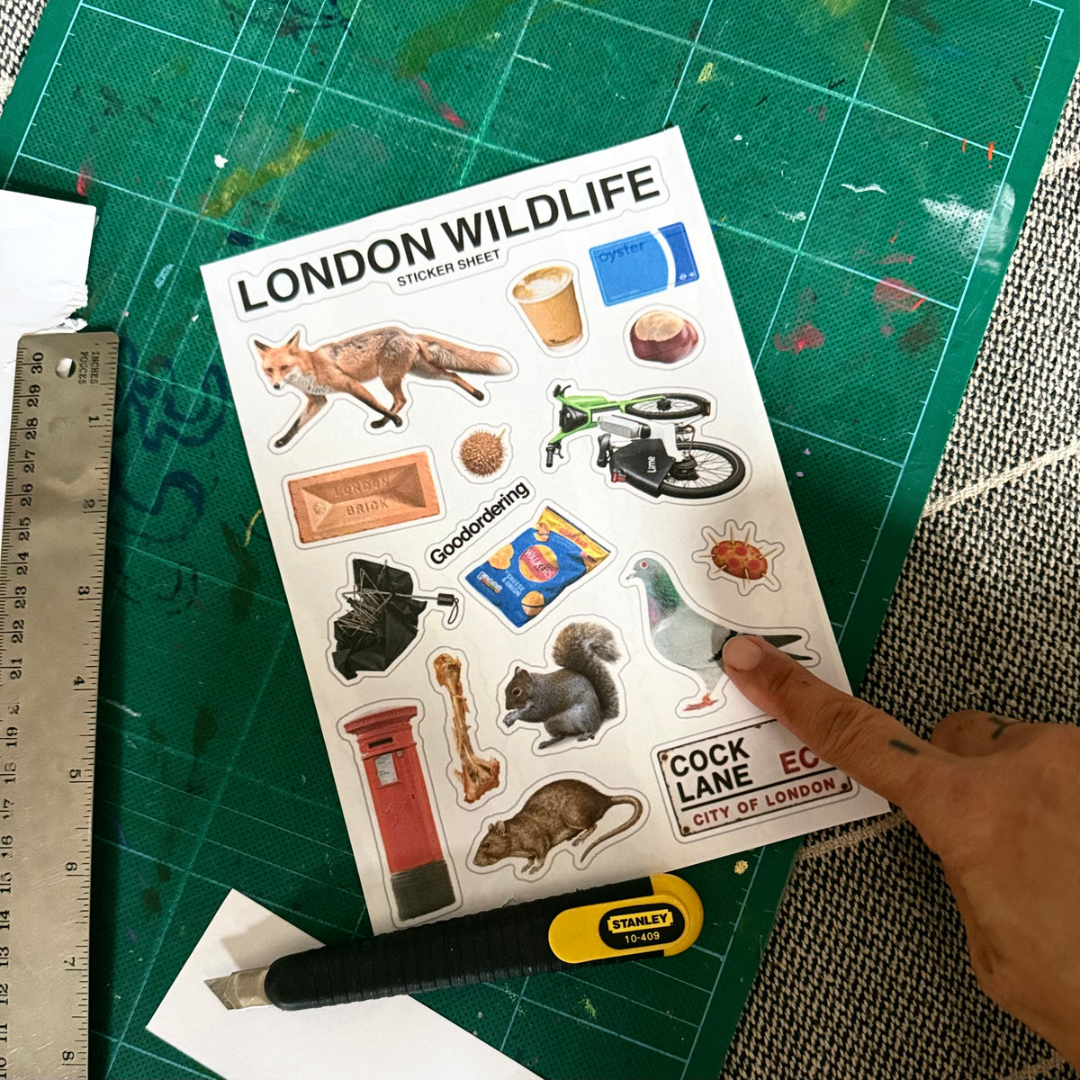 Goodordering 'London Wildlife Sticker Sheet' on a green cutting mat, surrounded by a ruler, utility knife, and textured surface.