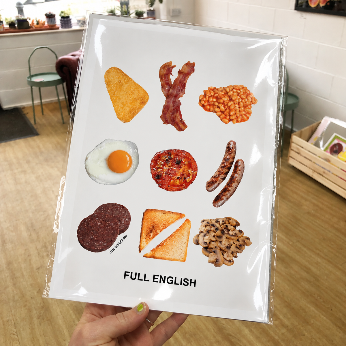 A hand holds a clear plastic-wrapped print featuring nine food illustrations arranged in a grid with the text 'FULL ENGLISH' at the bottom. The background shows a real indoor room with a wooden floor, chairs, and a basket.
