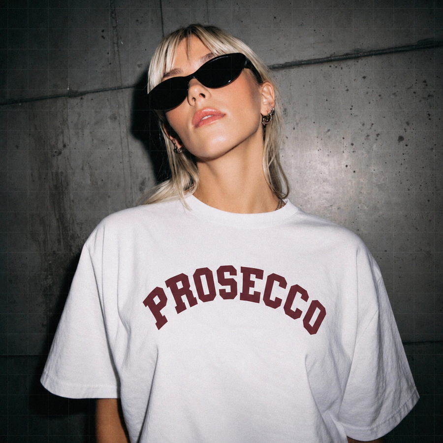 A blonde woman wearing black sunglasses and a white oversized t-shirt with 'PROSECCO' printed in dark red arched lettering, posing against a dark concrete wall.