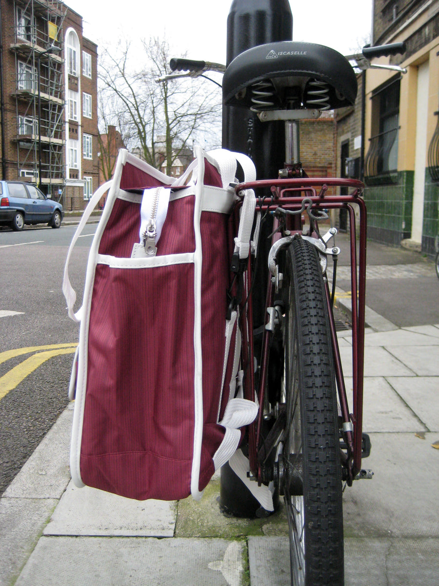 A burgundy and white bicycle pannier bag hanging from the rear rack of a dark red bicycle on a city sidewalk.