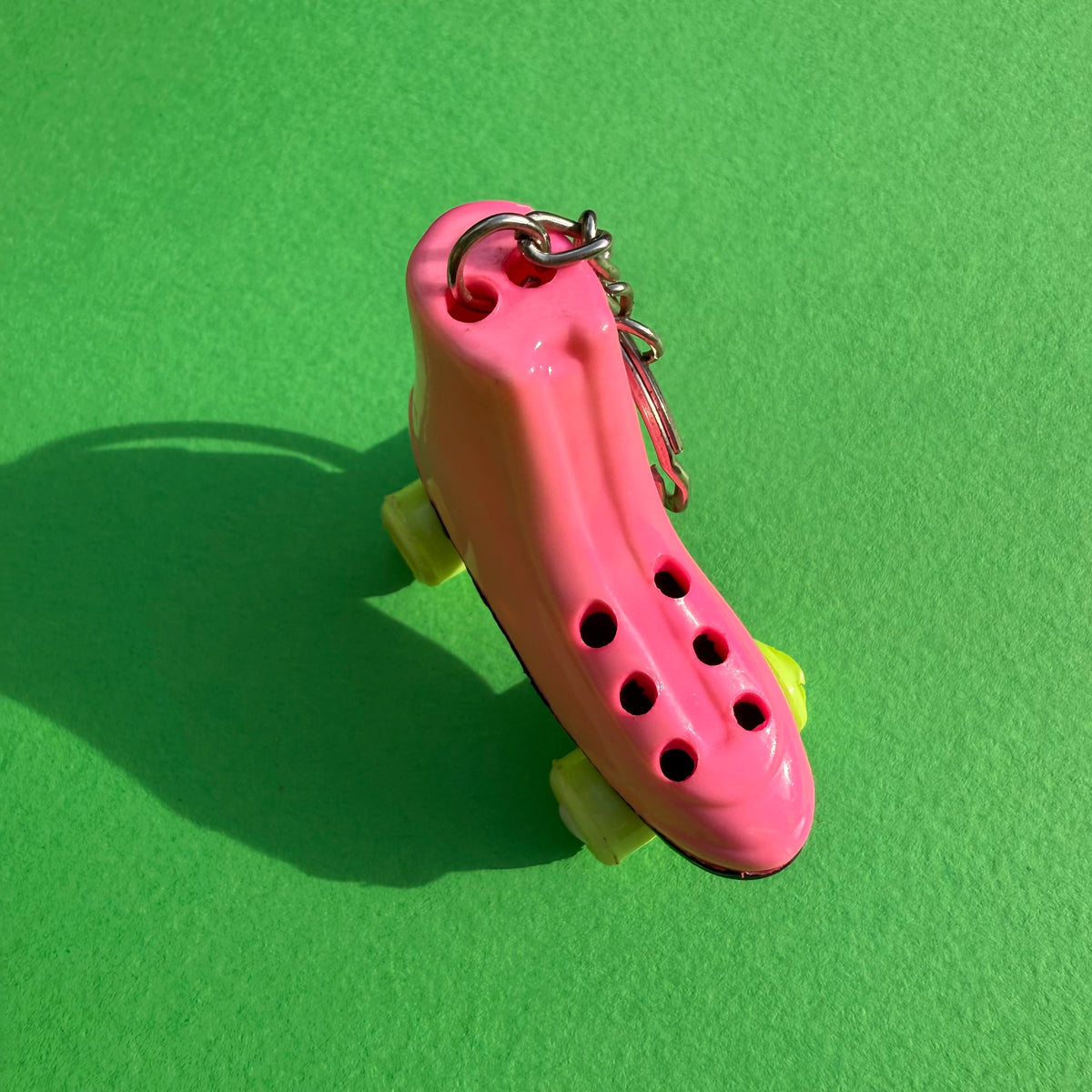 A novelty pink roller skate keychain with yellow wheels and a silver metal key ring, photographed on a bright green textured background.