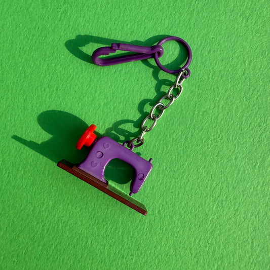 A miniature purple sewing machine keychain charm with a red wheel and brown base, attached to a silver metal chain and purple key ring, set against a bright green textured background.