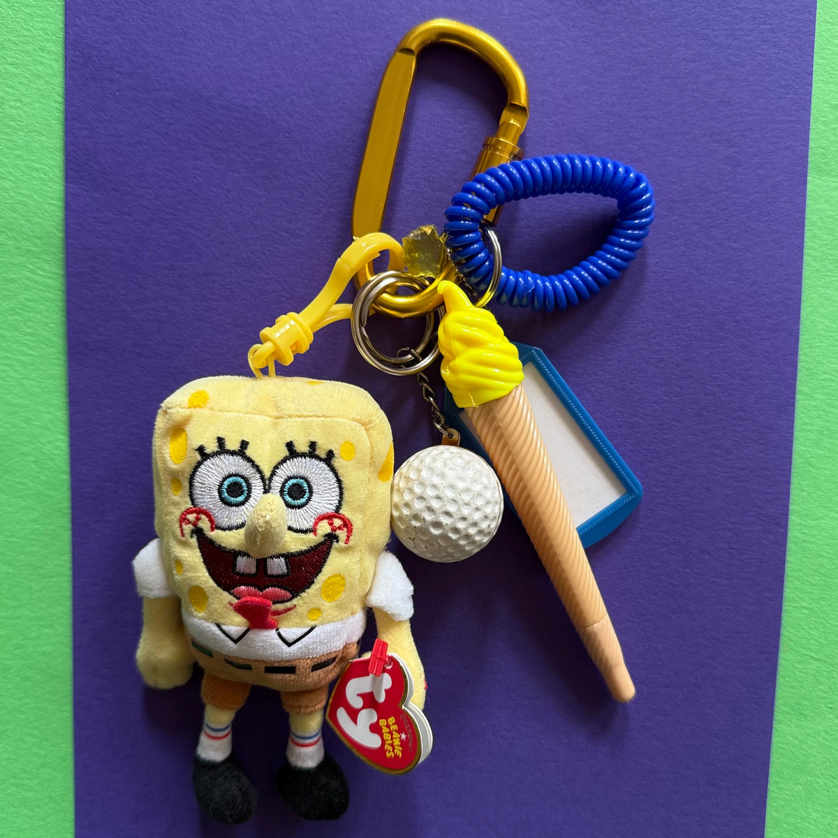 A SpongeBob-themed keychain set featuring a plush SpongeBob character, a yellow carabiner, a blue coiled strap, a white golf ball charm, a plastic cone-shaped accessory, and a red Tycho brand tag, displayed on a purple card against a green background.