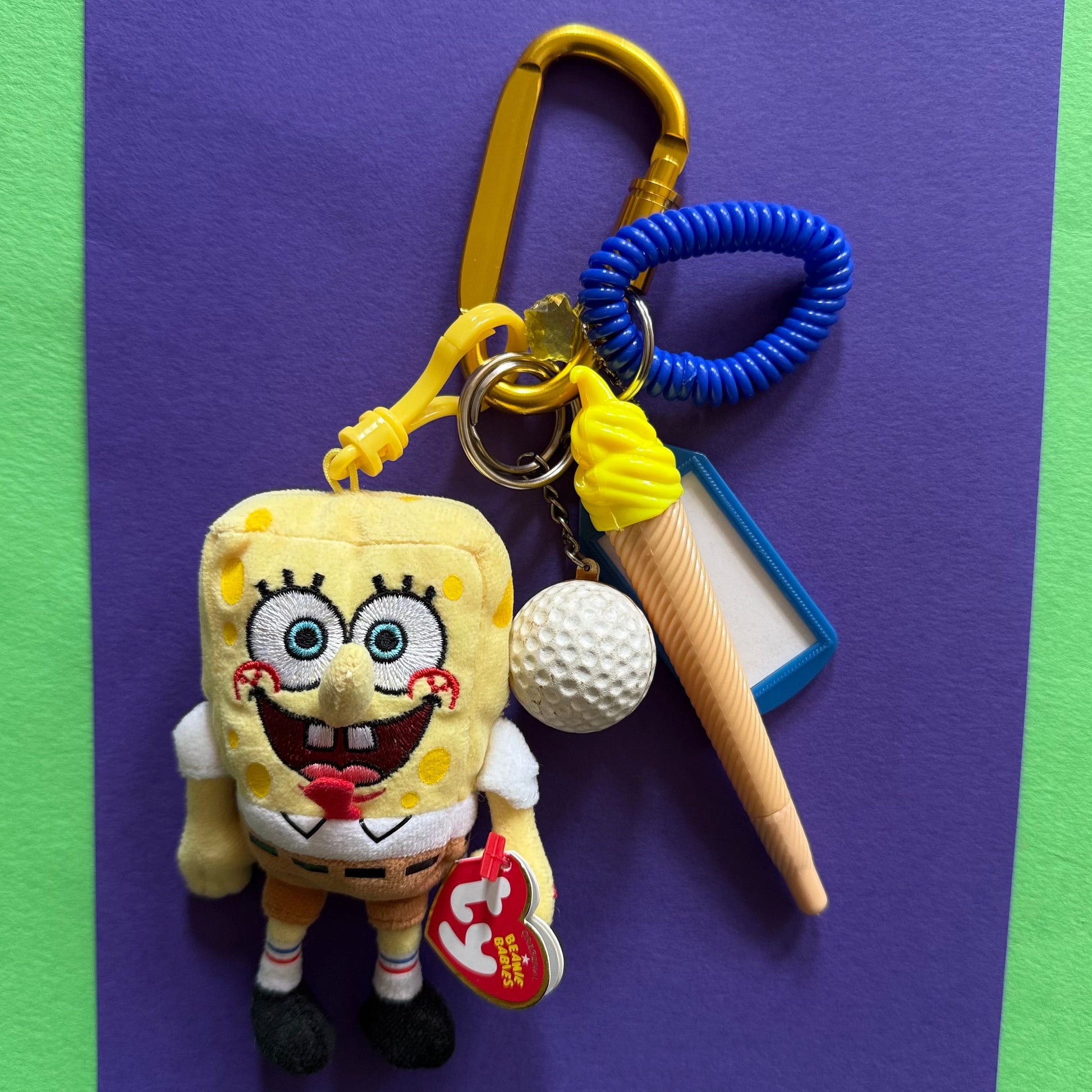 A SpongeBob-themed keychain set featuring a plush SpongeBob character, a yellow carabiner, a blue coiled strap, a white golf ball charm, a plastic cone-shaped accessory, and a red Tycho brand tag, displayed on a purple card against a green background.