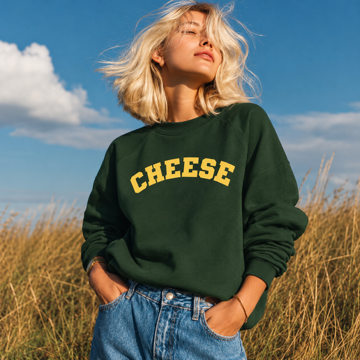 A woman outdoors in a field of dry grass wearing a dark green crewneck sweatshirt with the word 'CHEESE' printed across the chest in yellow varsity-style letters, paired with blue jeans under a blue sky with clouds.