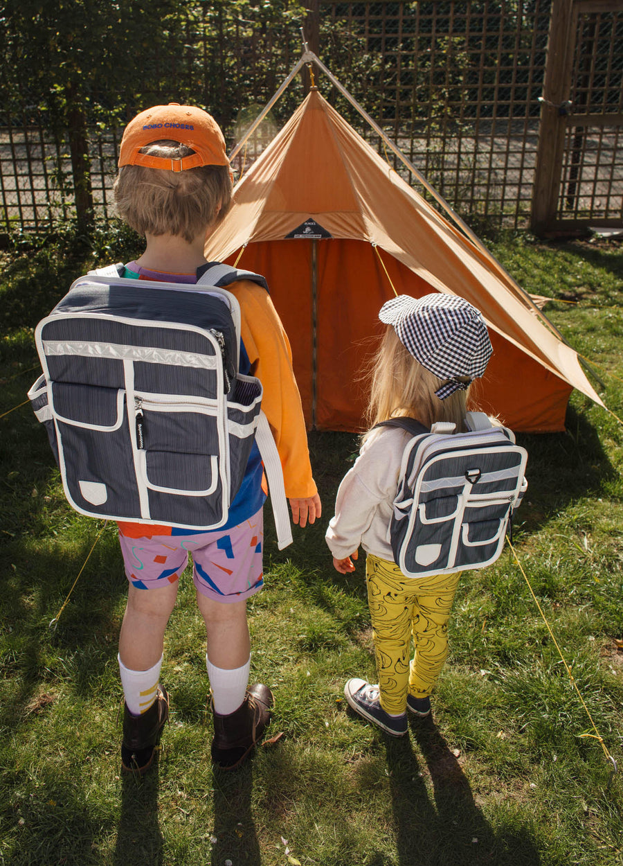 Two children seen from behind outdoors in front of an orange tent, each wearing a navy and white backpack. The child on the left wears an orange cap and patterned shorts, and the child on the right wears a blue-and-white checkered cap and yellow pants.