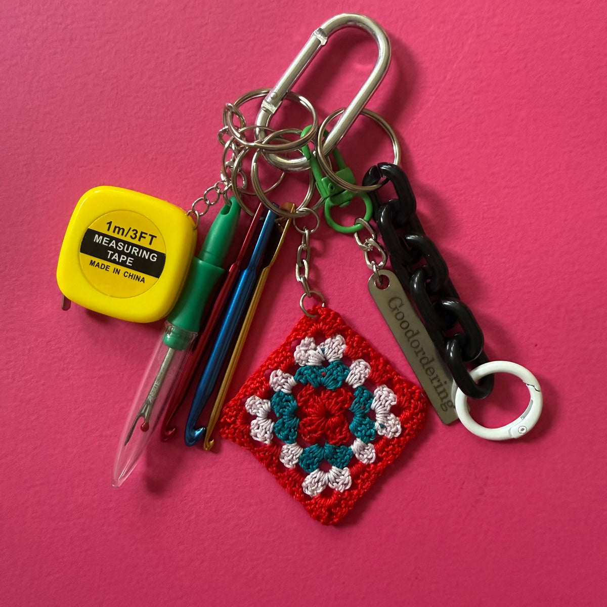 A crocheted granny square keychain charm in red, teal, and white, attached to a silver carabiner and keyring with pens, a measuring tape, and a black chain on a bright pink background. crochet-lover-bag-charm-keyring