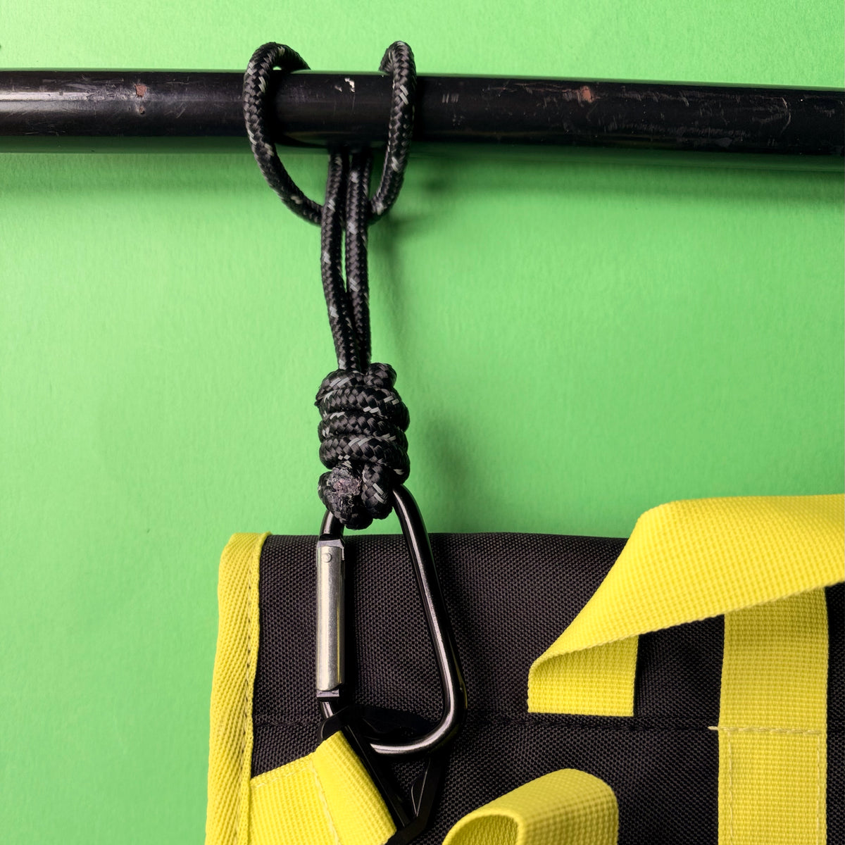 A close-up of a black paracord hanging loop and carabiner attached to a black metal bar, with a yellow and black bag visible below against a bright green background. goodordering-wheelchair-buggy-paracord-attachment-straps