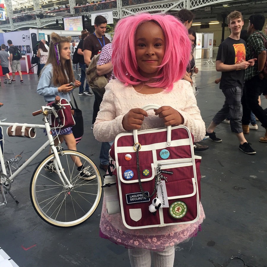 A young girl with bright pink hair stands in a crowded convention hall holding a burgundy and white goodordering backpack decorated with pins, stickers, and a 'Loving Richmond' patch. Other people and a bicycle are visible in the background.