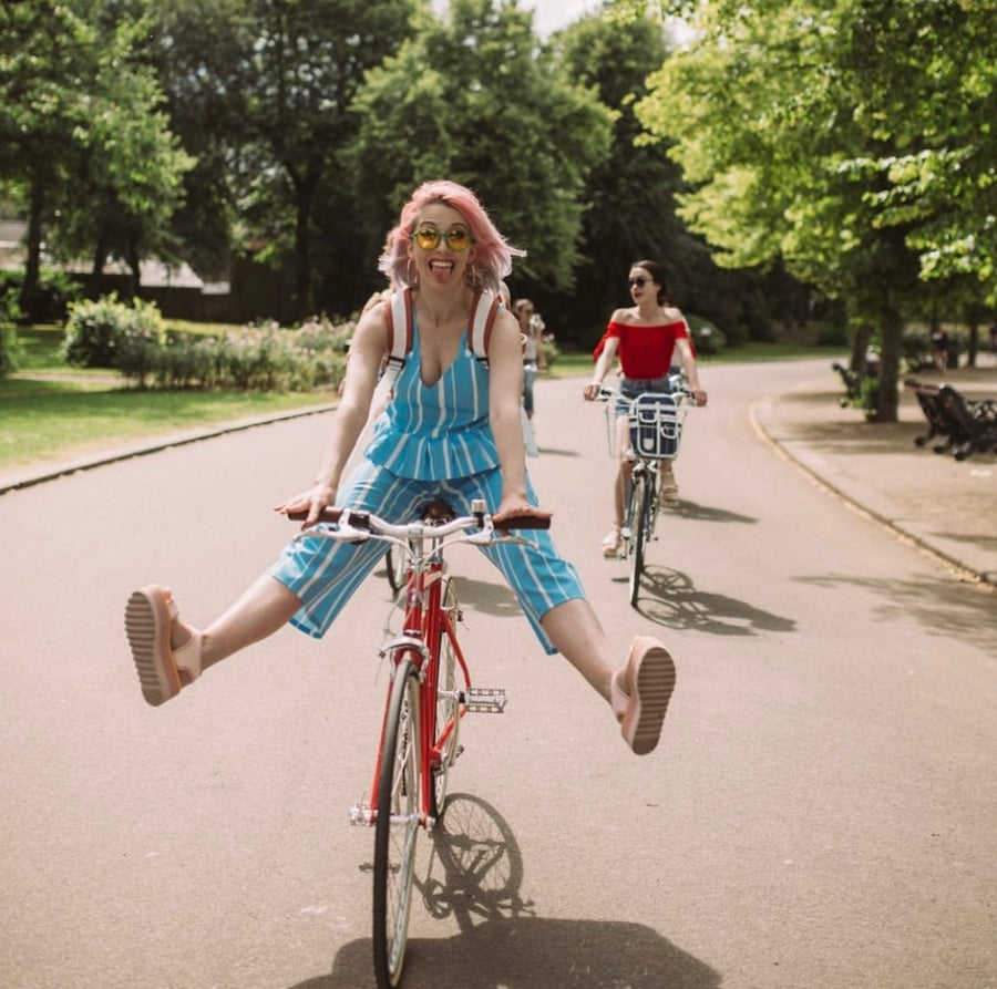 A woman with pink hair rides a red bicycle on a tree-lined path with her legs kicked up in the air, wearing a blue and white striped romper and sunglasses. Another woman on a bicycle follows behind her on the path.