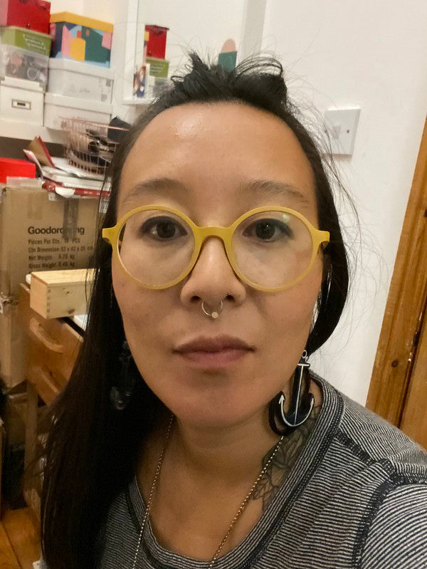 A close-up selfie of jacqui ma wearing round yellow goodordering eyeglasses, a nose ring, and a striped shirt, with a cluttered indoor room in the background.