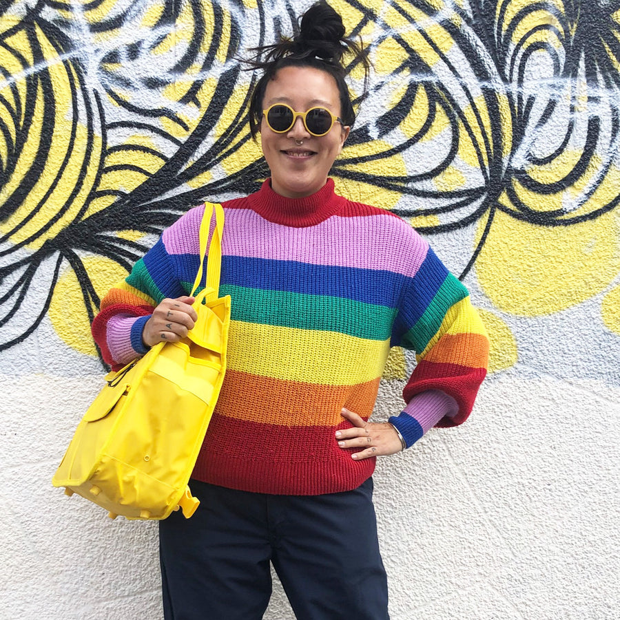 Goodordering founder Jacqui Ma wearing a vibrant rainbow-striped knit turtleneck sweater and round yellow-tinted sunglasses, holding a bright yellow tote bag against a textured wall with a yellow and black graffiti mural.