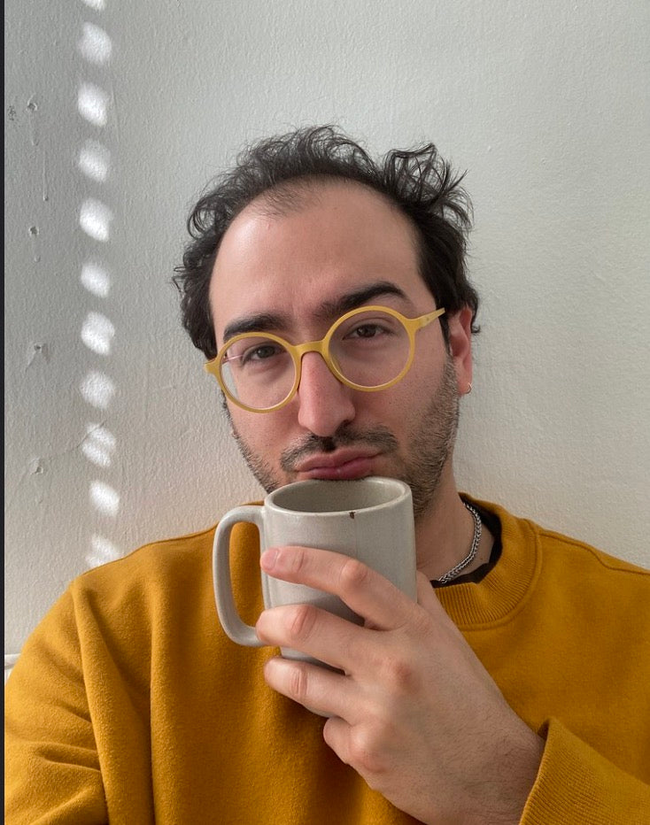 A man with dark hair and a beard wearing yellow round eyeglasses and a mustard yellow sweatshirt, holding a light gray ceramic mug against a white wall with dappled light.