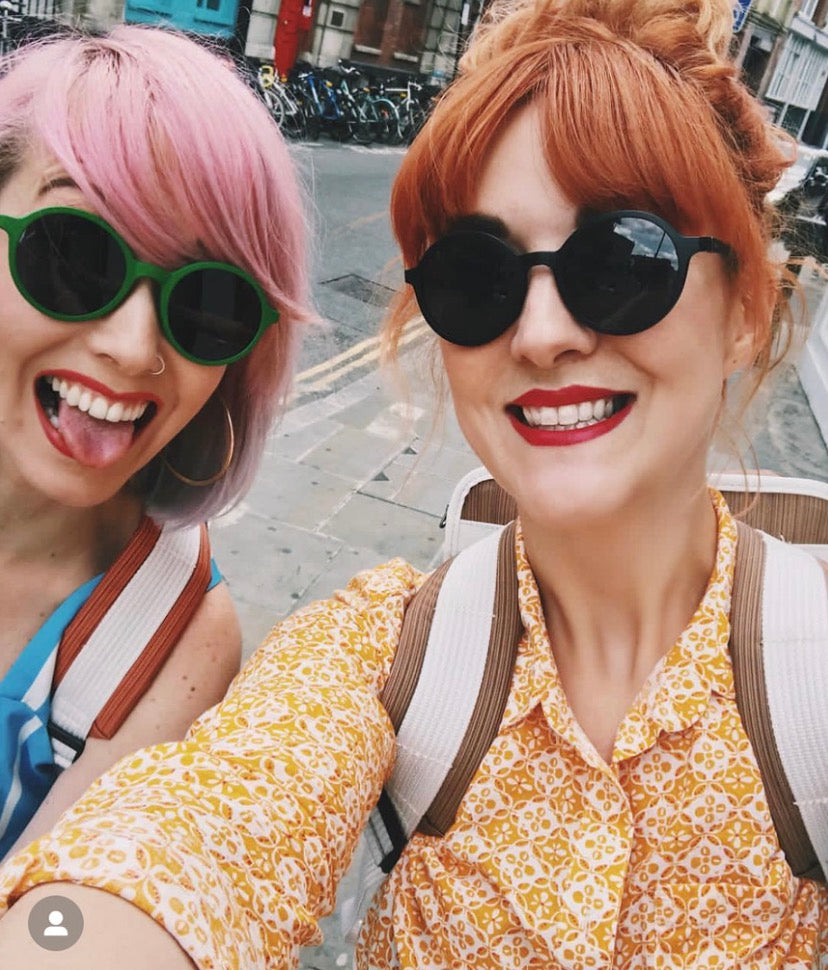 Two women taking a selfie outdoors on a city street. The woman on the left has pink hair and green sunglasses, while the woman on the right has red hair, black sunglasses, and a yellow patterned shirt.