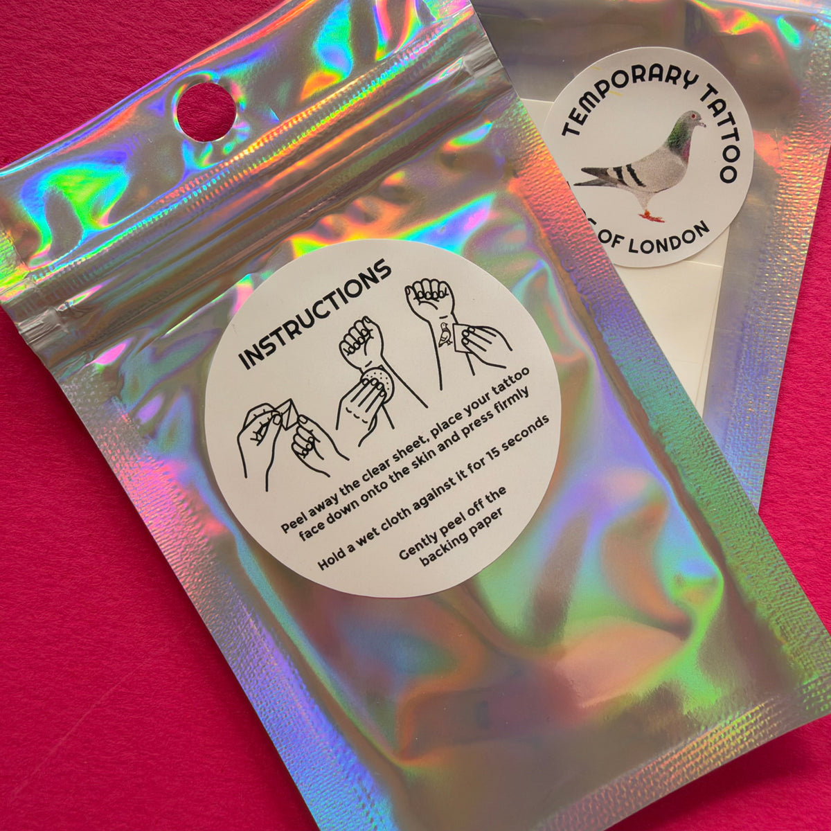 Two holographic zip pouches for temporary tattoos on a pink background. One pouch shows instructions with hand illustrations, and the other shows a round sticker that reads 'TEMPORARY TATTOO' and 'OF LONDON' with a pigeon logo.