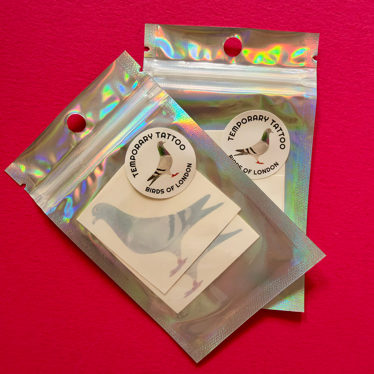 Two holographic zip-top pouches containing bird temporary tattoos on a bright pink background. The round white stickers read 'TEMPORARY TATTOO BIRDS OF LONDON'.