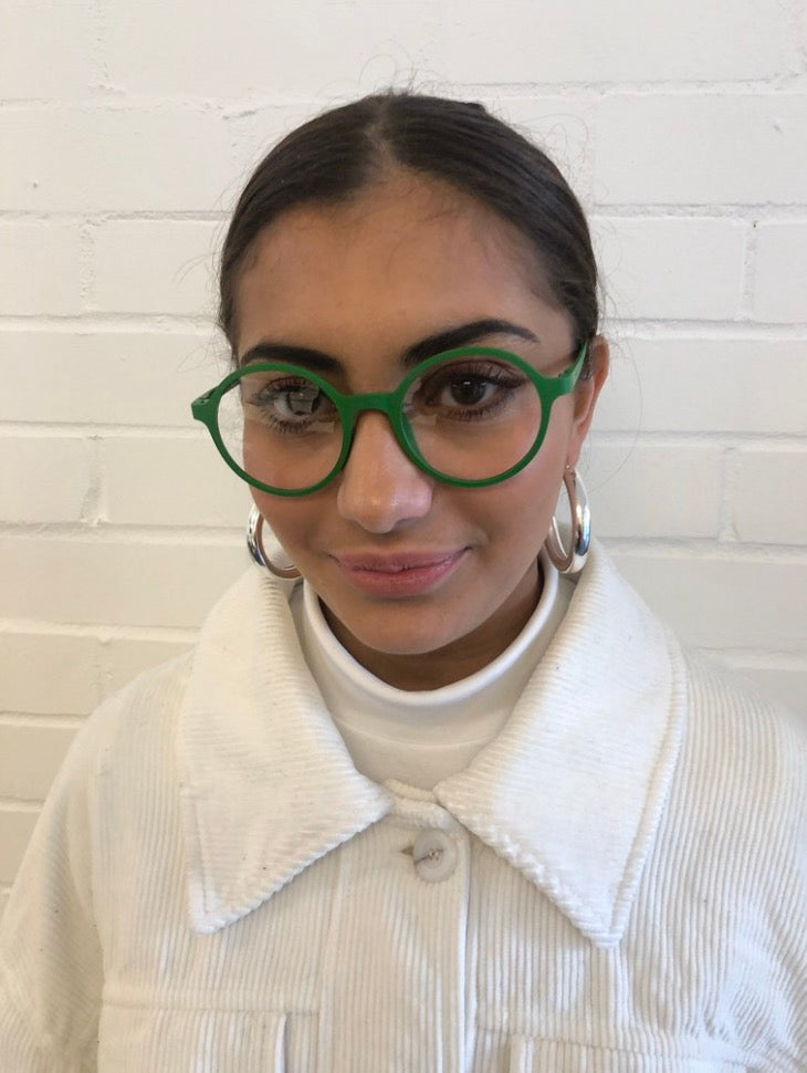 A woman with dark hair pulled back wears bright green round eyeglasses, silver hoop earrings, and a white ribbed turtleneck under a white corduroy jacket, standing against a white painted brick wall.