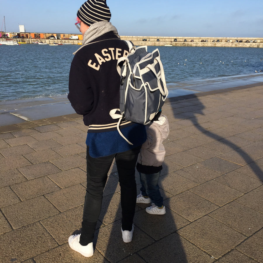 A person standing on a paved waterfront promenade, seen from behind, wearing a black varsity-style jacket with 'EASTER' in cream lettering on the back, a striped beanie, and carrying a gray and white Goodordering backpack. A child is partially visible beside them.