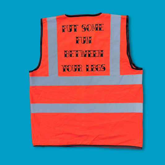 An orange high-visibility safety vest with reflective silver stripes, featuring the humorous text 'PUT SOME FUN BETWEEN YOUR LEGS' printed in black on the back, laid flat against a solid blue background.