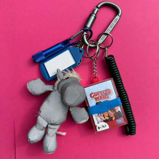A novelty keychain set featuring a gray plush donkey, a miniature Guess Who? box, a blue flashlight, a blue ID tag, and a black coiled cord, all attached to a silver carabiner on a bright pink background.