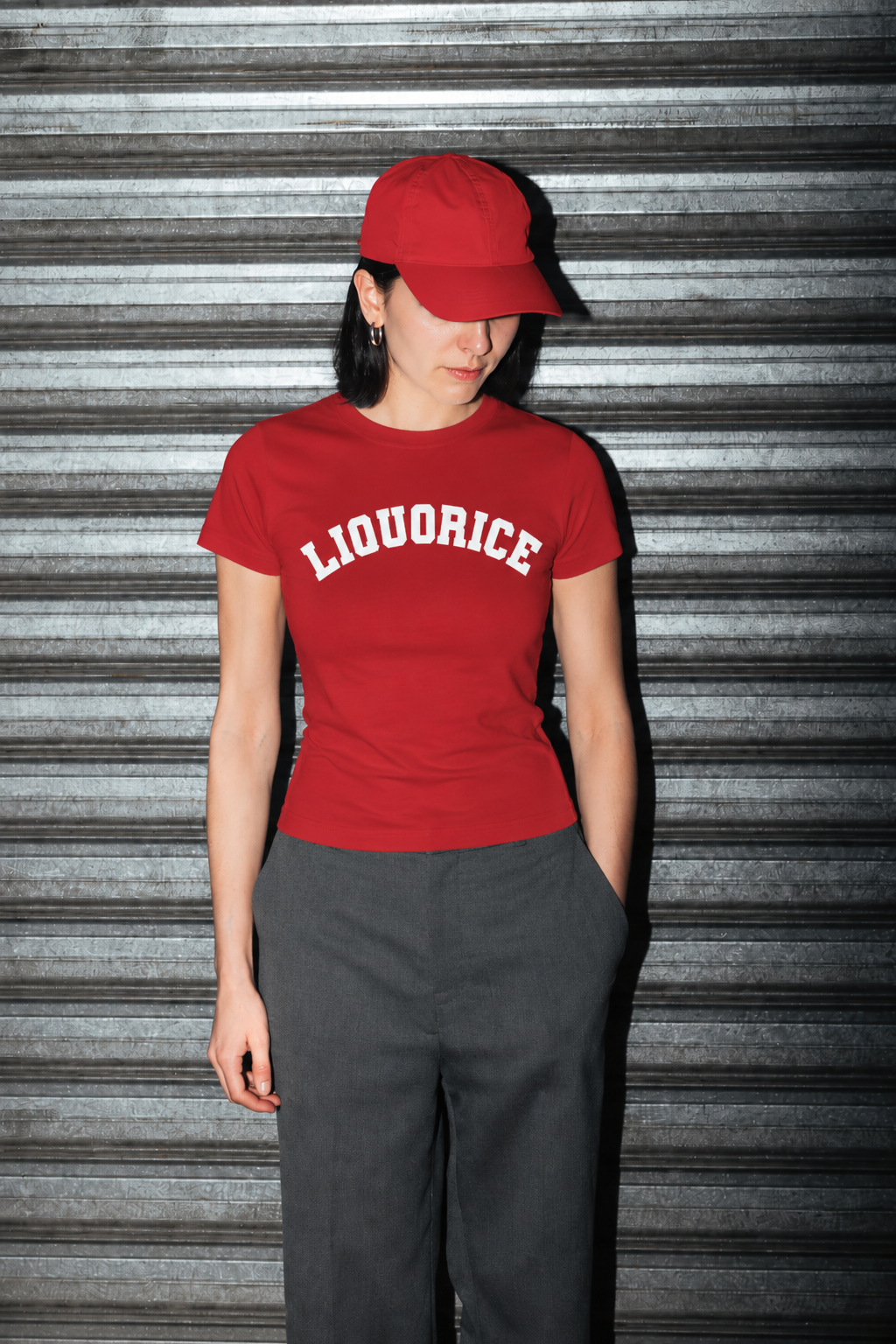 A woman wearing a red short-sleeve t-shirt with 'LIQUORICE' printed in white arched letters across the chest, paired with a matching red baseball cap and dark gray trousers, standing in front of a gray metal shutter.