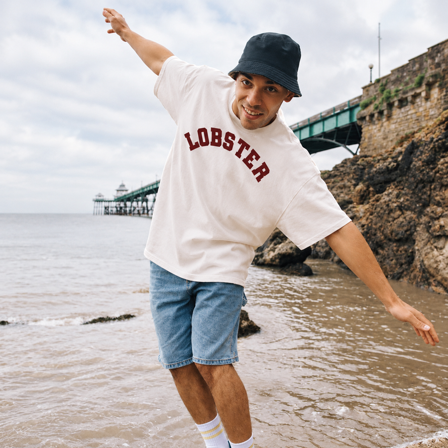 A man posing on a beach wearing a white t-shirt with 'LOBSTER' printed in red across the chest, blue denim shorts, a black bucket hat, and white socks, with a pier and rocky coastline in the background.