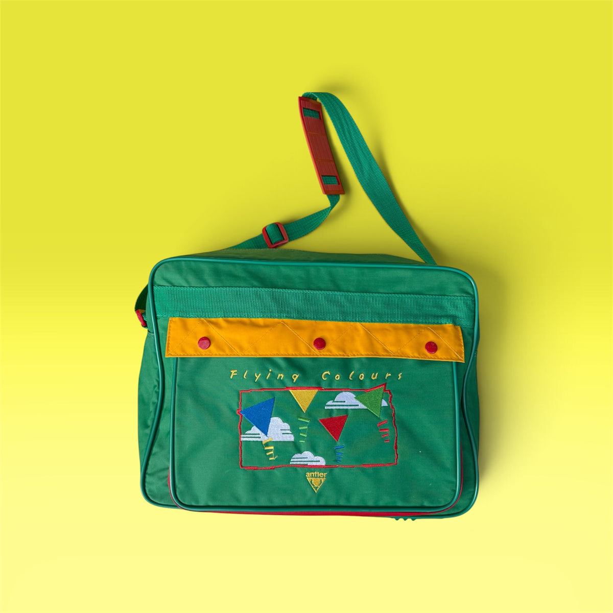 A green messenger bag with a yellow-orange horizontal stripe, red snap buttons, and a colorful embroidered patch on the front pocket, set against a solid yellow background.