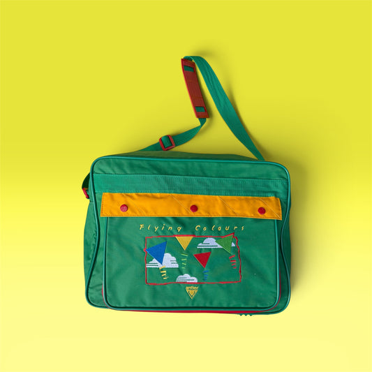 A green messenger bag with a yellow-orange horizontal stripe, red snap buttons, and a colorful embroidered patch on the front pocket, set against a solid yellow background.
