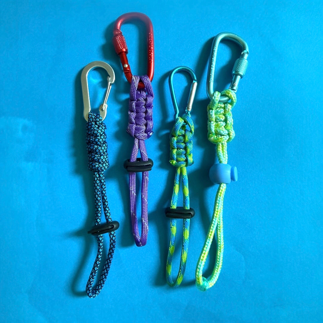 Four paracord keychains with carabiners in blue, purple, green, and light blue, arranged vertically on a blue background.