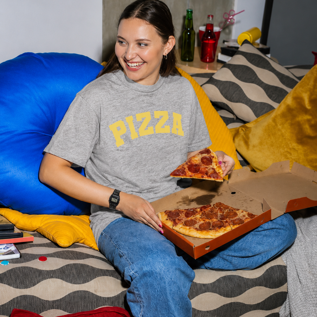 A woman sitting on a couch in a cozy living room setting, wearing a gray t-shirt with 'PIZZA' printed in yellow varsity letters. She is holding a slice of pepperoni pizza in one hand and a whole pizza box in the other, surrounded by snacks, pillows, and drinks.