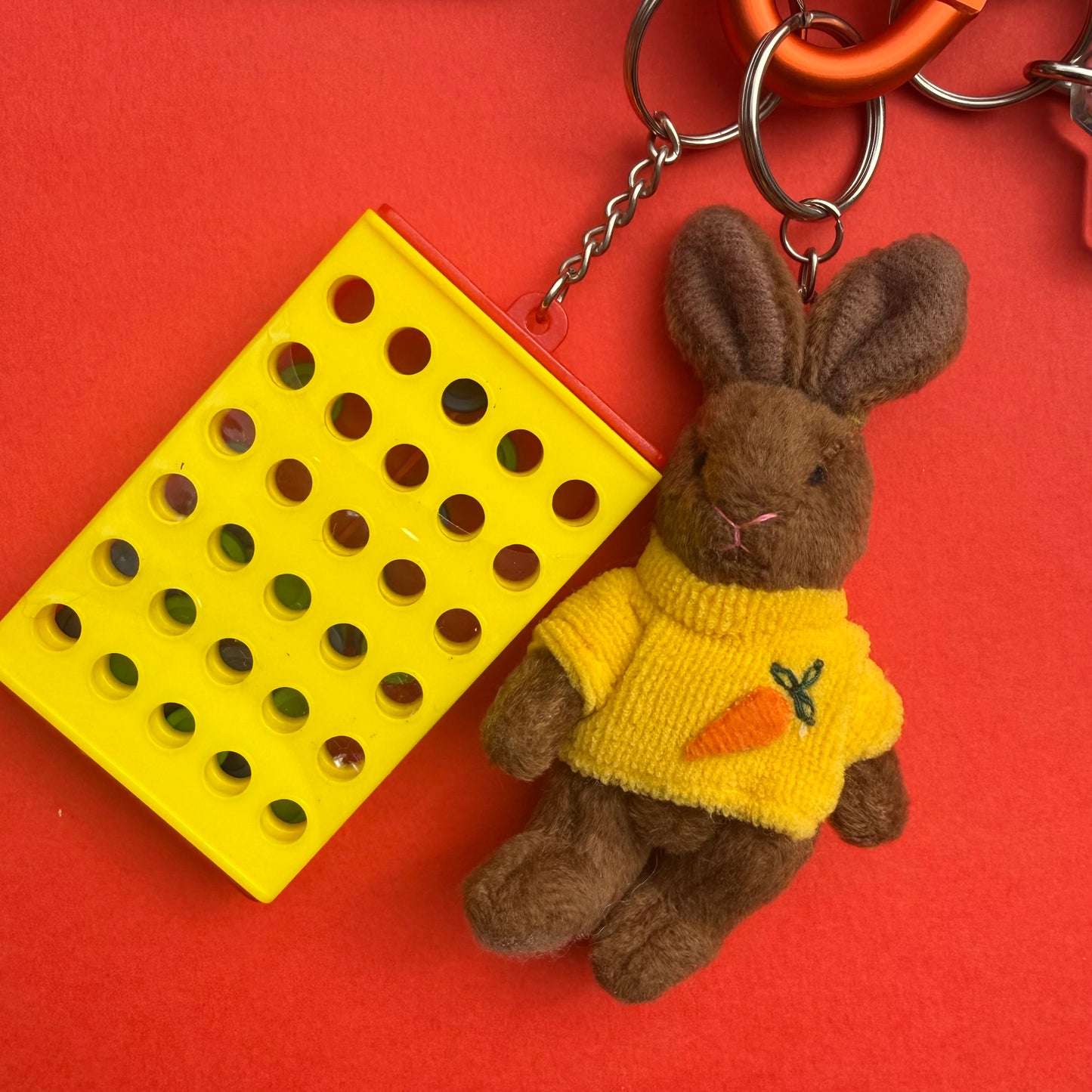 A plush brown bunny keychain wearing a yellow sweater with a carrot motif, attached to metal rings, next to a bright yellow rectangular plastic item with circular cutouts, all on a solid red background.