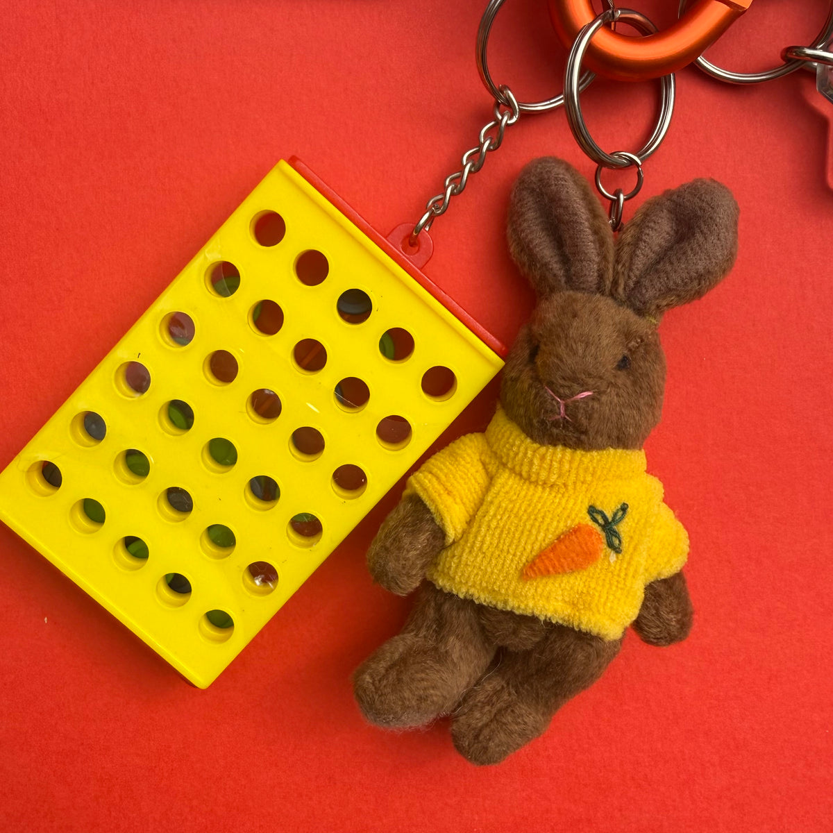 A plush brown bunny keychain wearing a yellow sweater with a carrot motif, attached to metal rings, next to a bright yellow rectangular plastic item with circular cutouts, all on a solid red background.