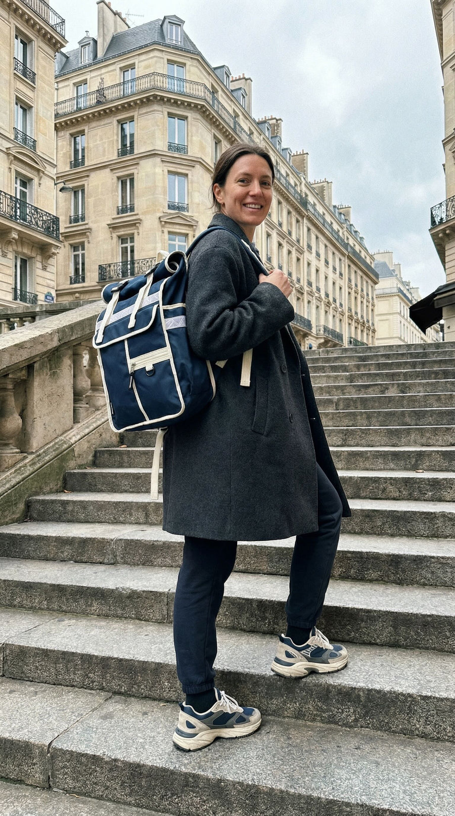 A woman stands on outdoor stone stairs in front of Parisian-style buildings, wearing a dark coat, dark pants, and sneakers, and carrying a blue and white backpack.