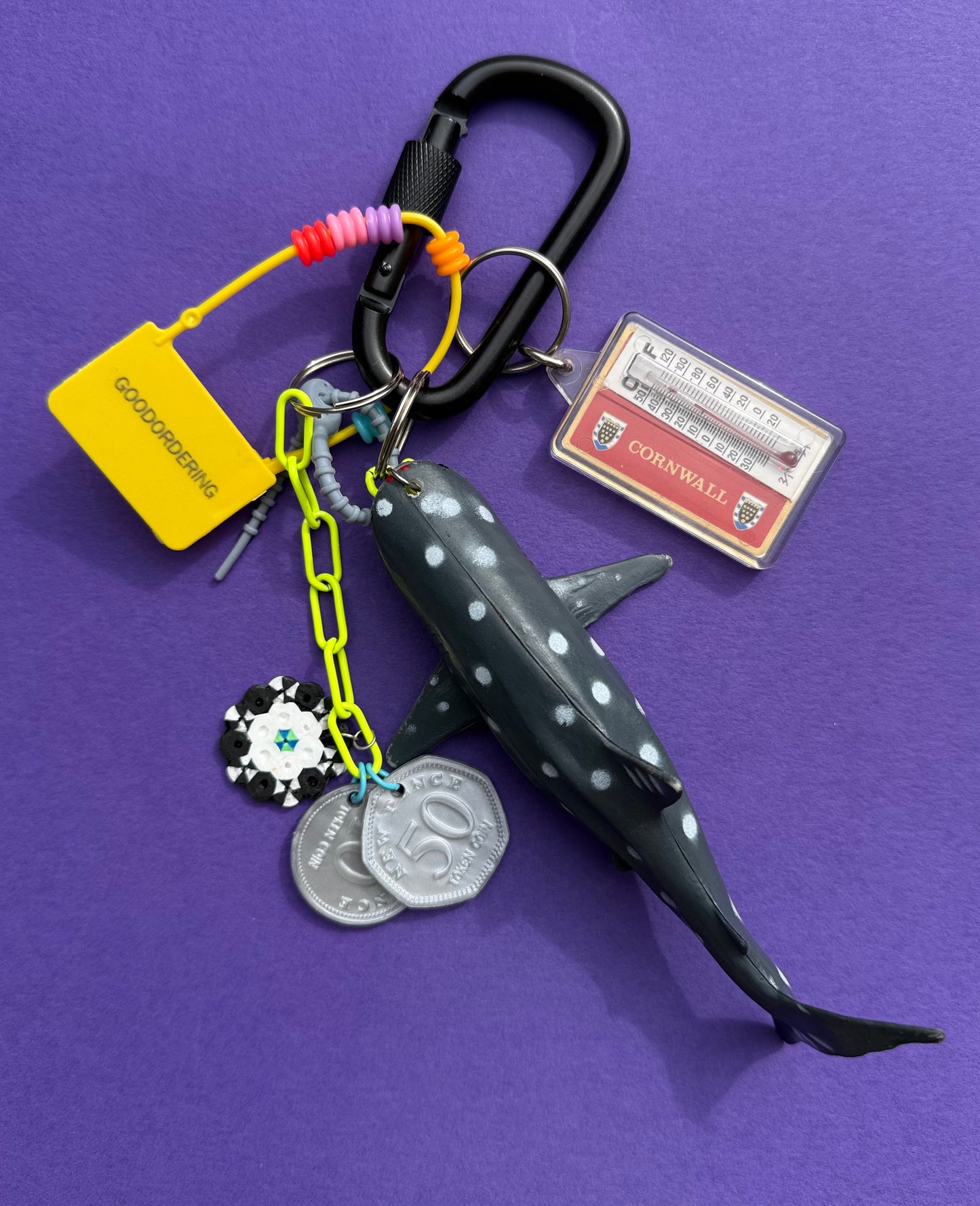 A novelty keychain featuring a shark-shaped charm with white spots, attached to a black carabiner, neon yellow chain, silver coin charms, a yellow 'GODOPRIORISING' tag, and a Colorado Avalanche collectible sticker, all displayed on a solid purple background.