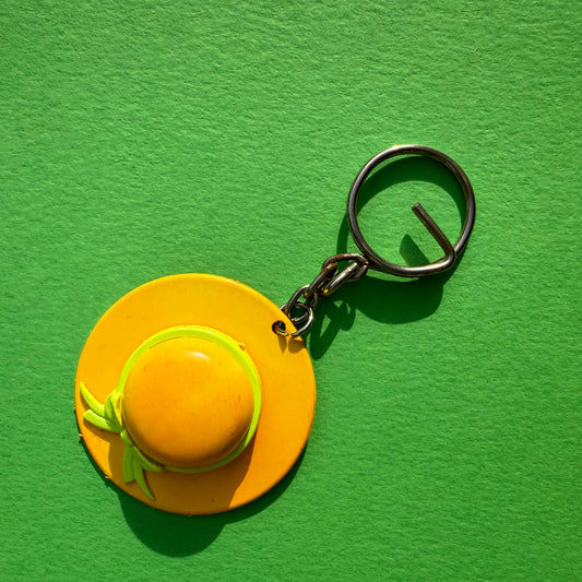 A novelty keychain shaped like a small yellow sun hat with a green band and bow, attached to a black split ring, set against a textured bright green background.