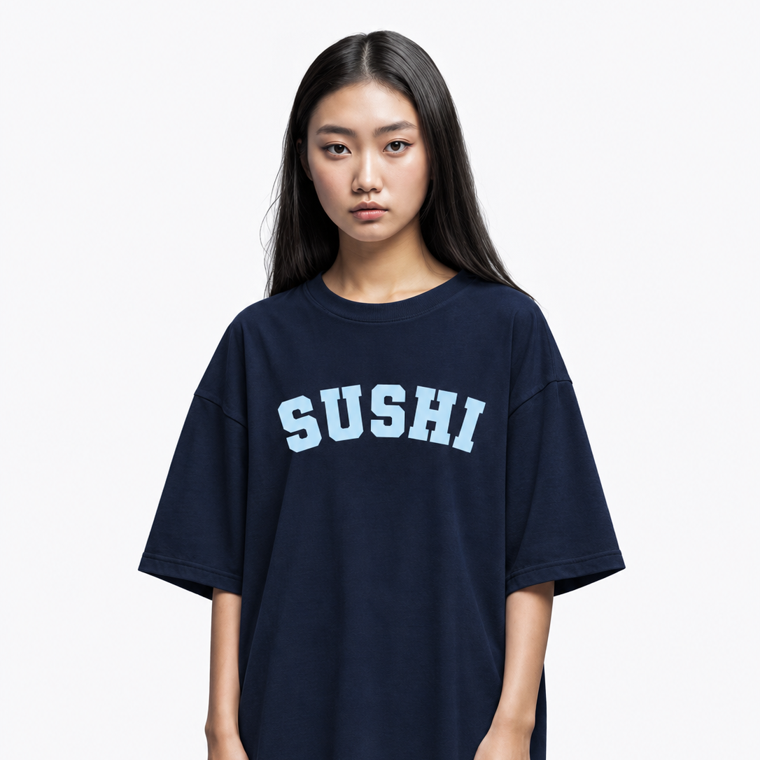 A young woman modeling an oversized dark navy blue t-shirt with the word 'SUSHI' printed across the chest in light blue varsity-style lettering, set against a plain light gray background.