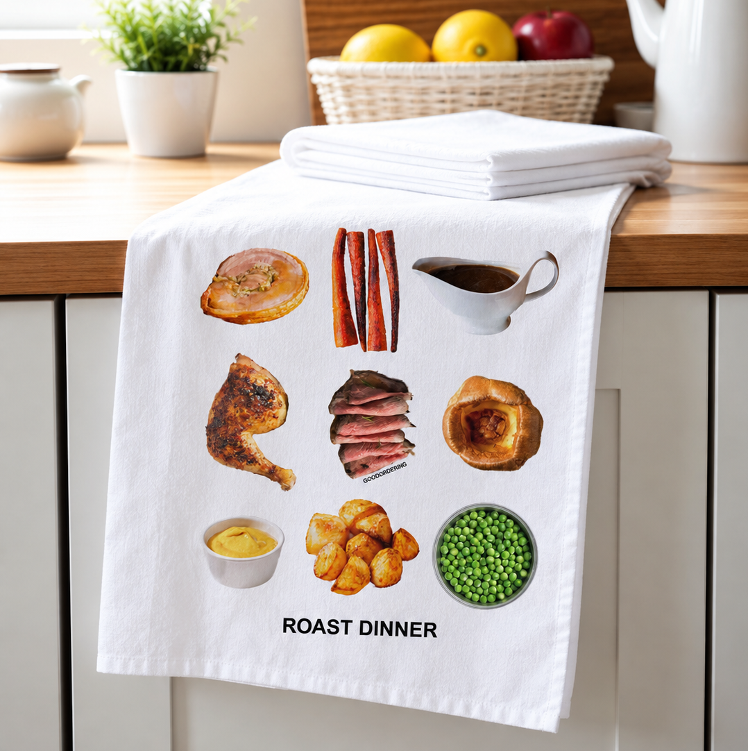 A white kitchen towel draped over a wooden countertop, printed with a roast dinner theme featuring food illustrations and the text 'ROAST DINNER'. The background shows a softly blurred kitchen setting with a basket of lemons and a teapot.
