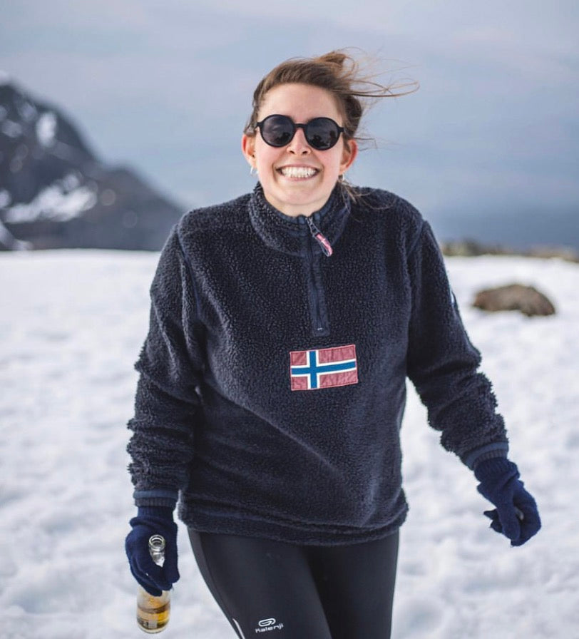 A woman smiling in a snowy outdoor setting, wearing a dark navy fleece quarter-zip pullover with a Norwegian flag patch on the chest, black leggings, and goodordering magnetic sunglasses.